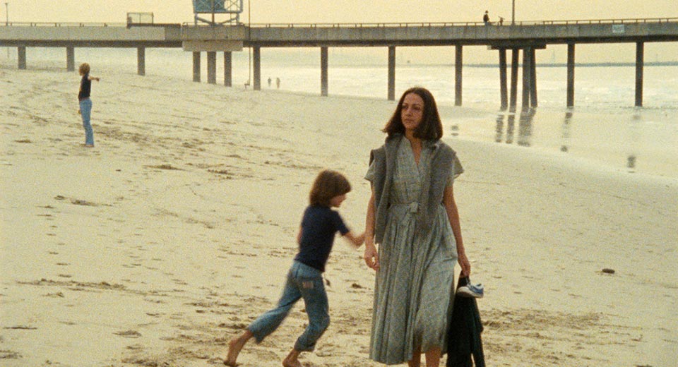 a woman walks along a beach holding her son's jacket, who runs freely behind her in the setting sun