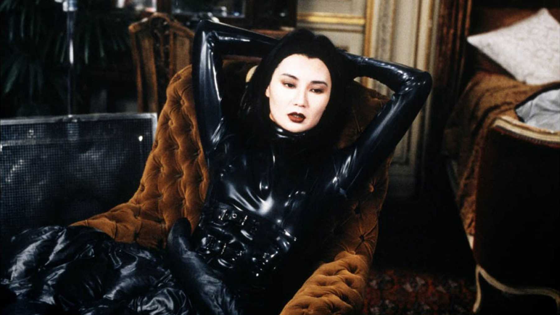 a woman in a leather catsuit lounges in an orange chair with her hands behind her head