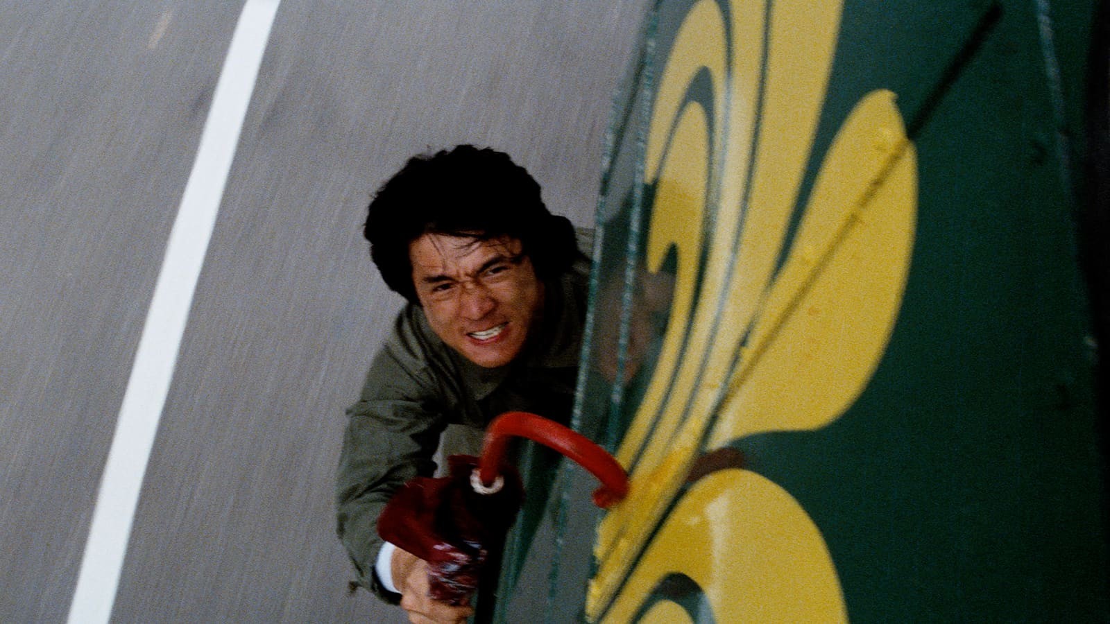 a shaggy haired man clings, grimacing, to a moving vehicle with only an umbrella