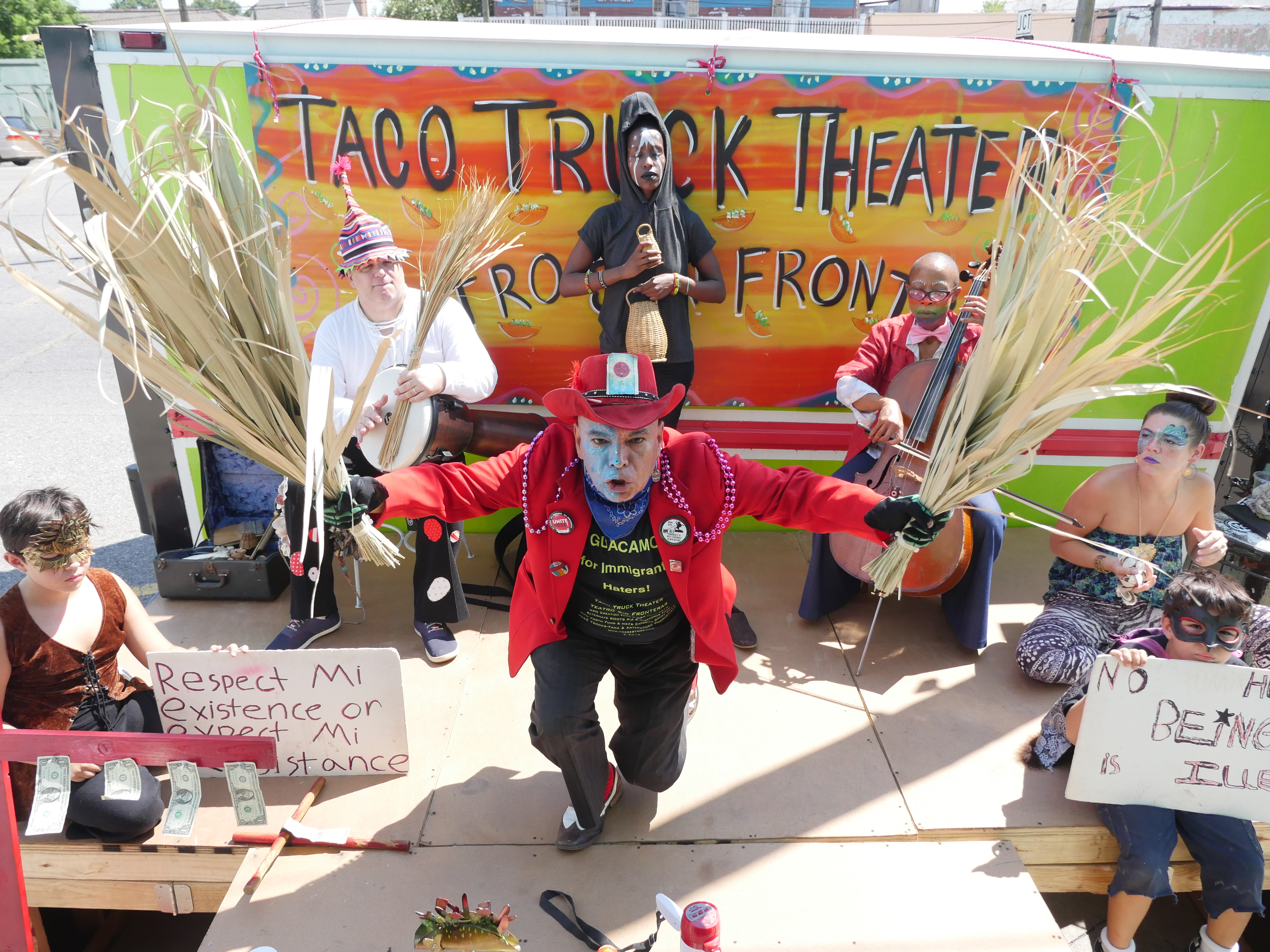 a man in bright red jacket and hat holds palm fronds in both hands as a crew of performers circles around him 