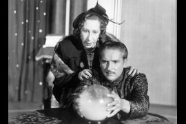 a man gazes intently into a crystal ball while a woman places her hands on her shoulders and stares likewise