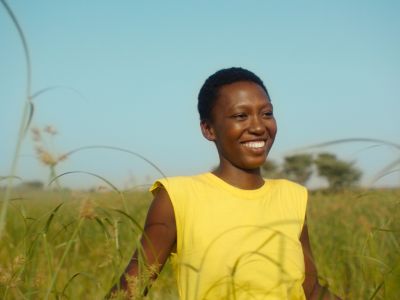 A woman wearing a bright yellow tunic smiles in a grassy field in a still from Banel & Adama